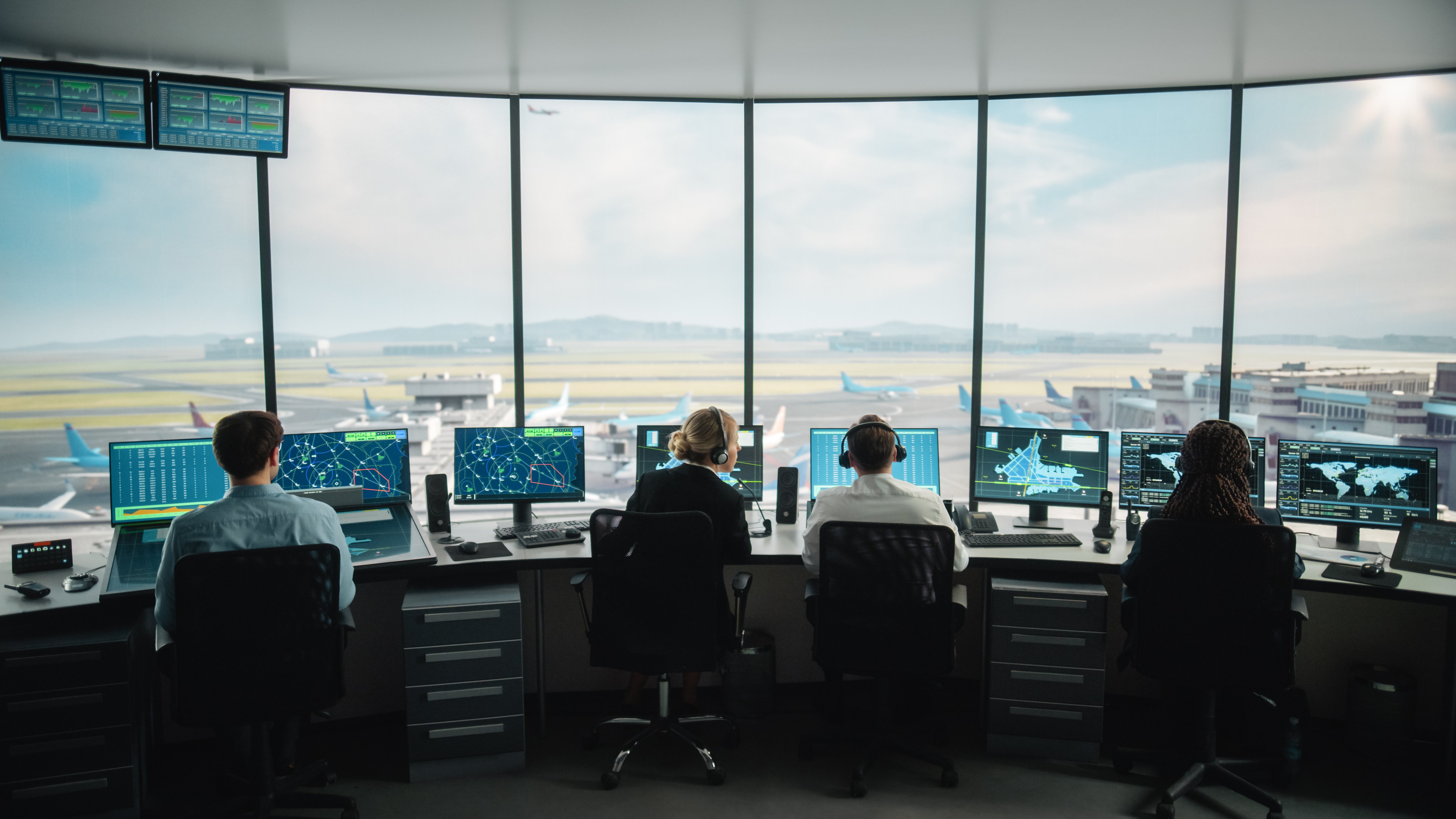 Quatre contrôleurs aériens sont assis à des postes de travail dans la tour de contrôle d'un aéroport, chacun surveillant plusieurs écrans affichant des radars, des trajectoires de vol, des cartes de l'aéroport et des données globales. De grandes fenêtres panoramiques offrent une vue claire de la piste de l'aéroport, des avions qui roulent et des bâtiments du terminal. L'équipe coordonne les mouvements des avions dans un environnement sécurisé et de haute technologie