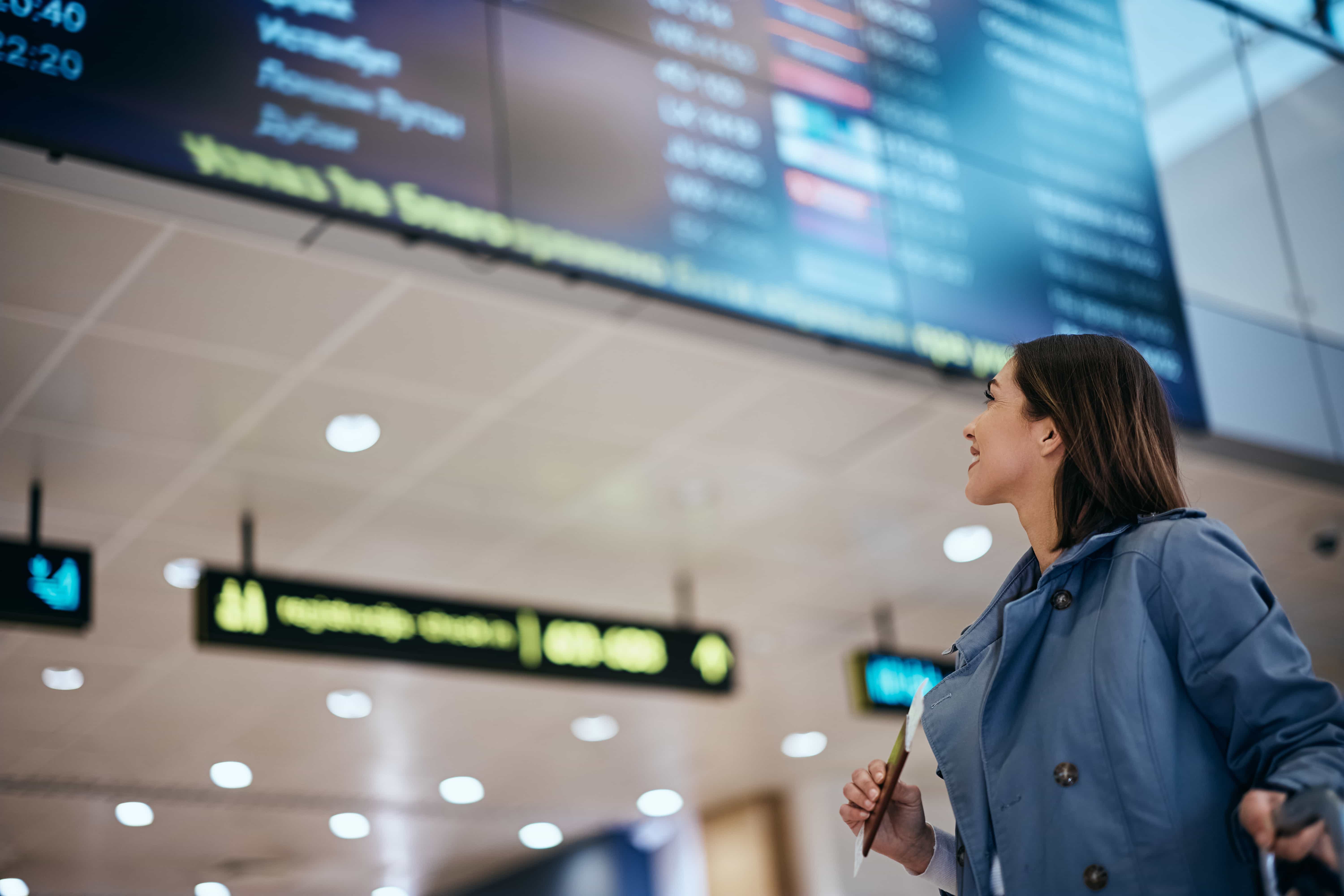  Une femme vêtue d'une veste bleu clair et d'une chemise blanche regarde un grand tableau des départs dans un aéroport. Elle sourit légèrement et tient ce qui semble être un passeport ou un billet dans sa main droite. Le tableau des départs, qui n'est pas net, affiche diverses informations sur les vols. Le plafond de l'aéroport avec ses lumières est également visible en arrière-plan.