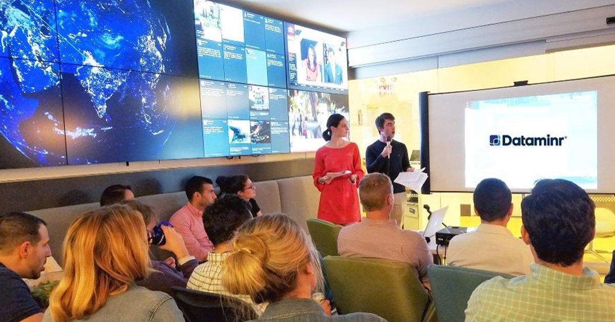 Des employés de Dataminr dans une salle de réunion, assis en face d'un projecteur et à côté d'un mur vidéo présentant les médias sociaux et les actualités.