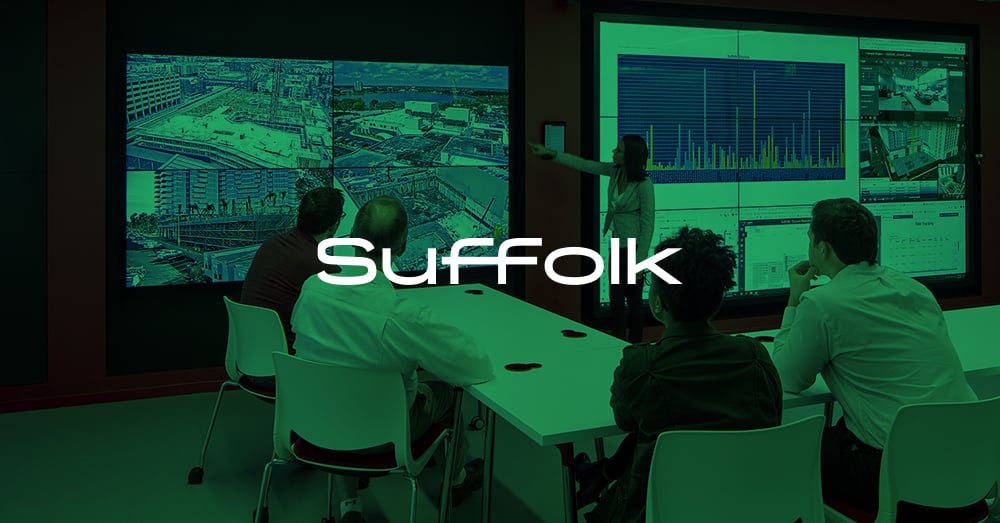 Un employé debout fait une présentation à l'aide d'un mur vidéo, 4 employés regardent la présentation assis à une table dans le L.A. Smart Lab de Suffolk avec une superposition verte et un logo.