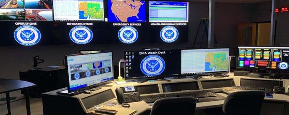 Le centre de contrôle vide de l'EMA avec des postes de travail et un mur vidéo affichant des photos, des données et des cartes.