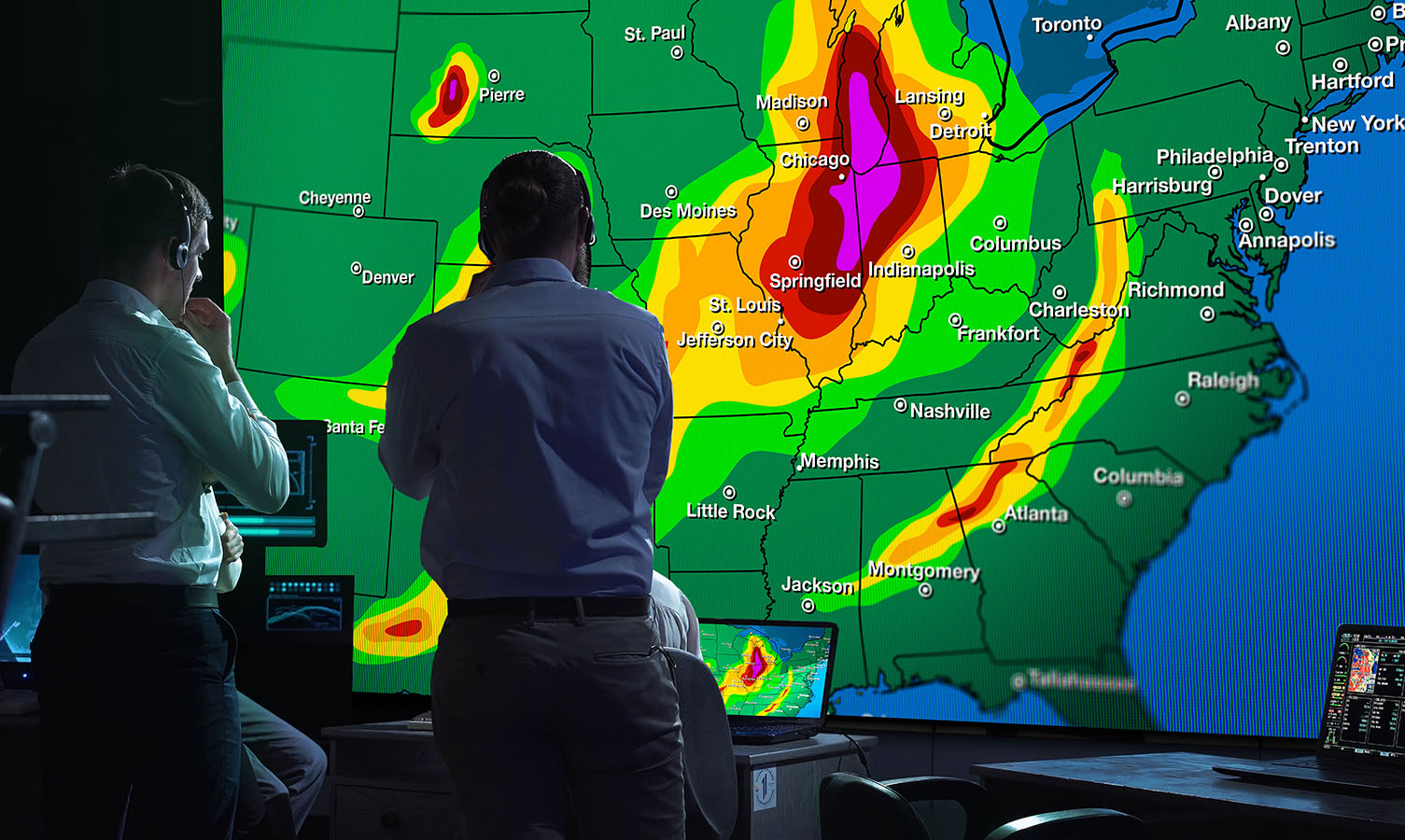 2 hommes dans une salle de contrôle de la météo, regardant un grand mur vidéo affichant une carte météorologique des États-Unis.