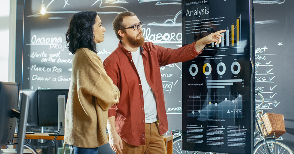 Un homme et une femme discutent d'un tableau de bord de données sur un écran de télévision, avec un tableau noir avec les détails du projet derrière eux.