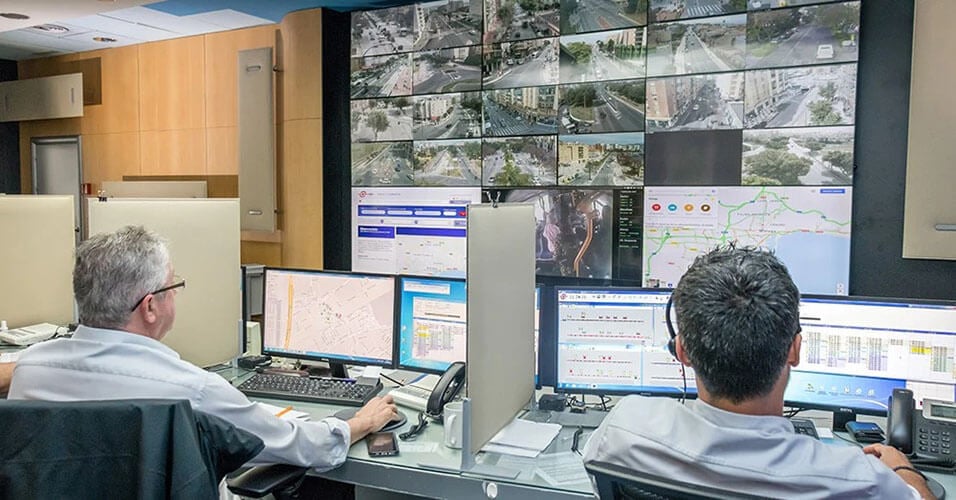 Deux employés de l'EMT surveillent les opérations de transport en commun à partir de leur poste de travail et d'un mur vidéo affichant des images de caméra en direct, des cartes de transport en commun et des sites Web.