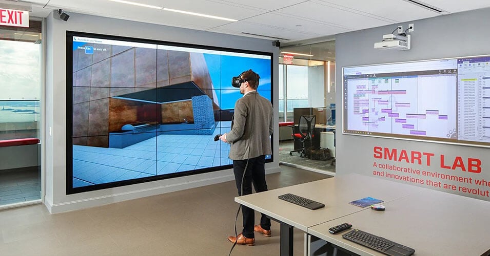 La salle du Smart Lab de Suffolk avec un bureau et un homme qui explore la réalité virtuelle, et un mur vidéo qui affiche ce qu'il voit virtuellement.