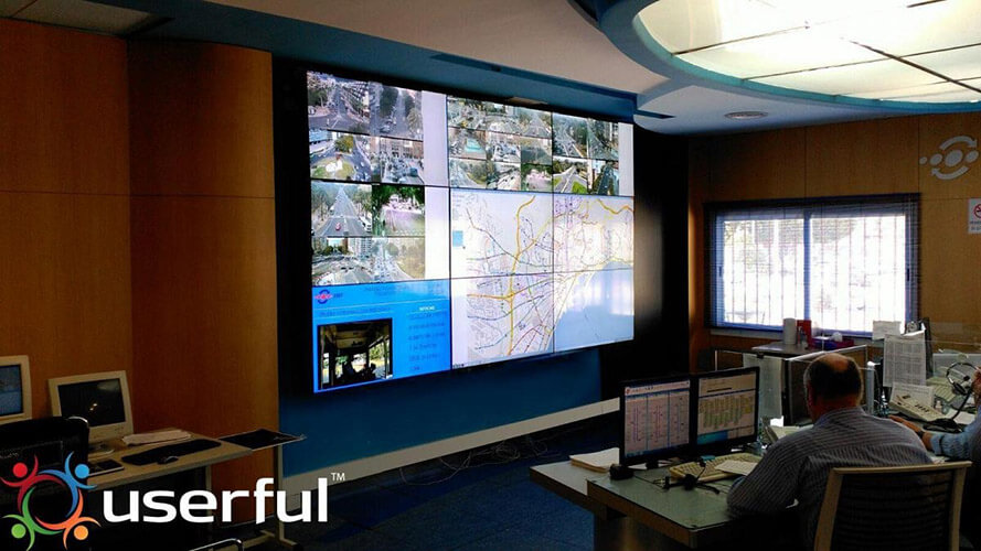 Deux employés de l'EMT surveillent les opérations de transport en commun à partir de leur poste de travail et d'un mur vidéo affichant des images de caméra en direct, des cartes de transport en commun et des sites Web.